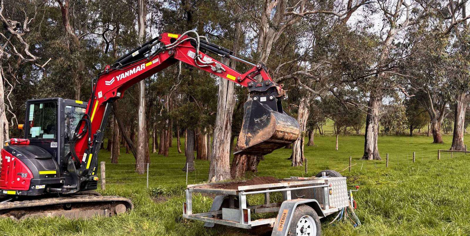 A Yanmar ViO55 excavator lifting a bucket of soil over a tandem trailer in a stand of eucalypts on grazing land in regional Victoria.